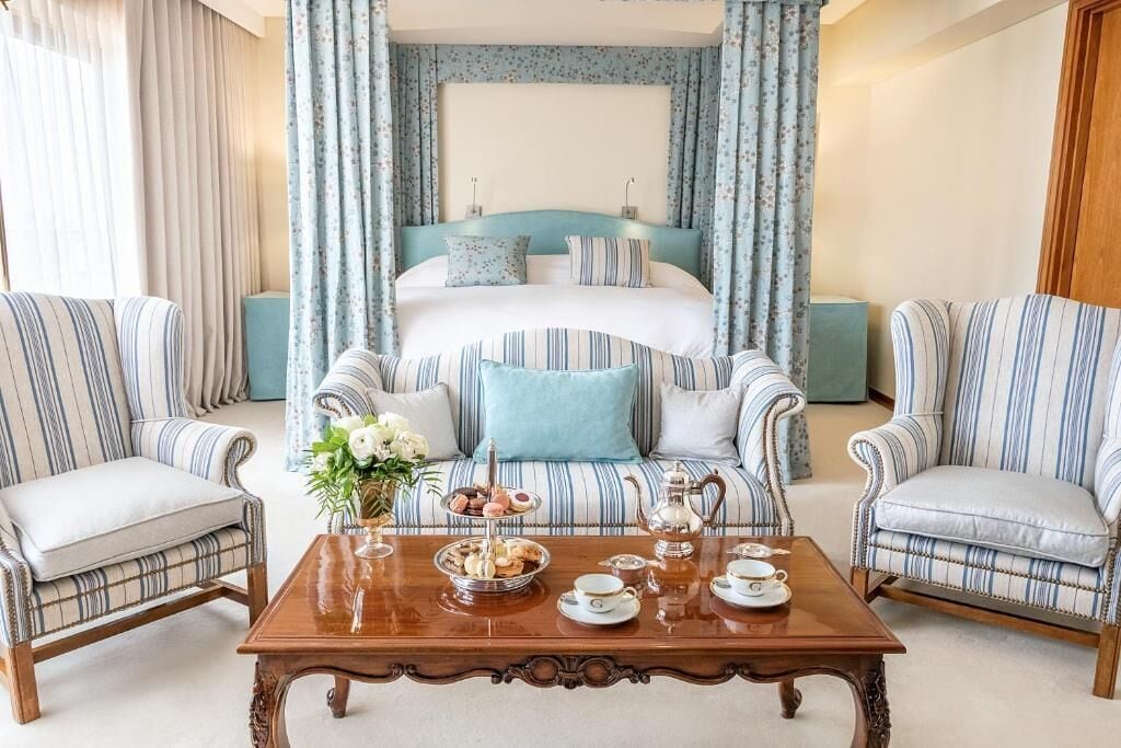Habitación de hotel con una cama grande, dos sillones, una mesita de centro con juego de té y cortinas.