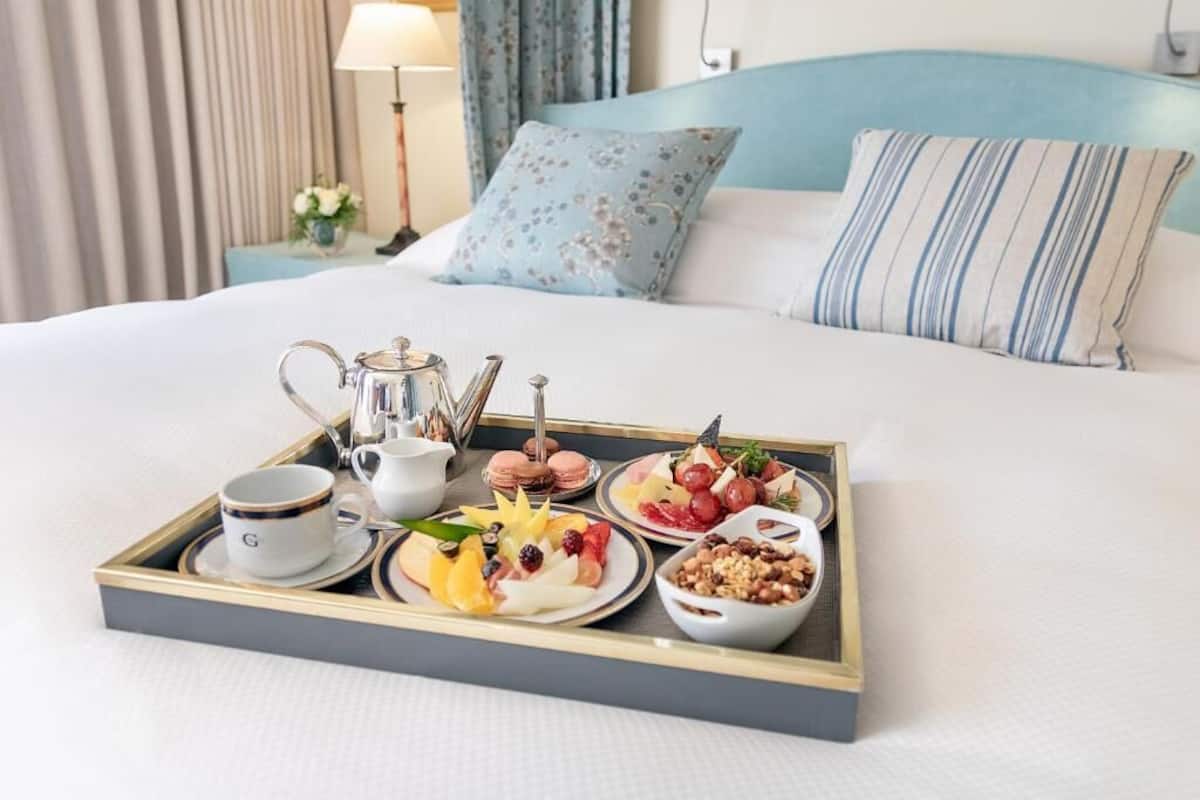 Een keurig opgemaakt ontbijtkarretje op een bed met een bloemenkussen en een gestreept kussen, een lamp en gordijnen op de achtergrond.