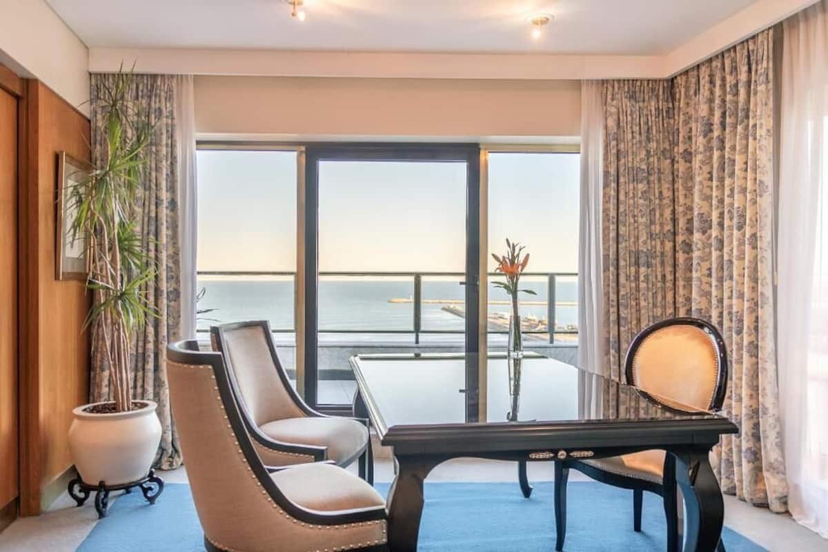 Een hotelkamer met balkon, bureau, twee stoelen, een potplant en uitzicht op zee.