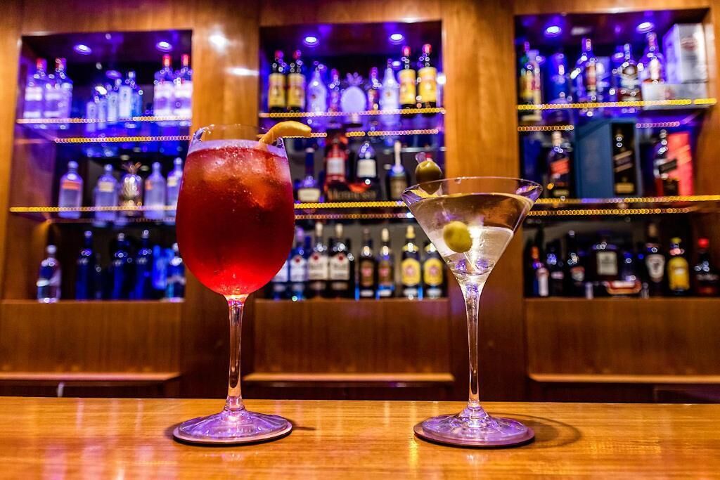 Two cocktails on a bar counter with shelves of liquor bottles in the background.