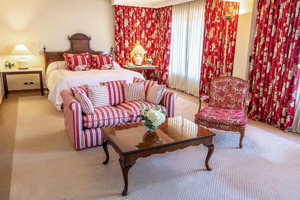 A hotel room with a bed, a sofa, a chair, a nightstand, a lamp, and curtains.