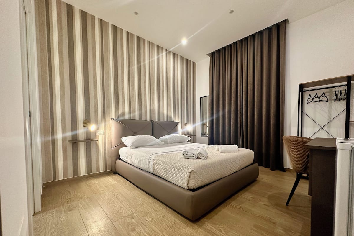 A modern hotel room with a large bed, wooden flooring, striped wallpaper, and a desk with a chair.