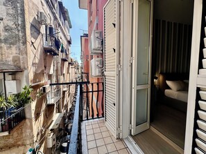 A narrow balcony with a view of adjacent buildings, a sliding glass door leading to a bedroom with a bed and a nightstand, and an air conditioning unit on the wall. - Casa Morelli (Naples)