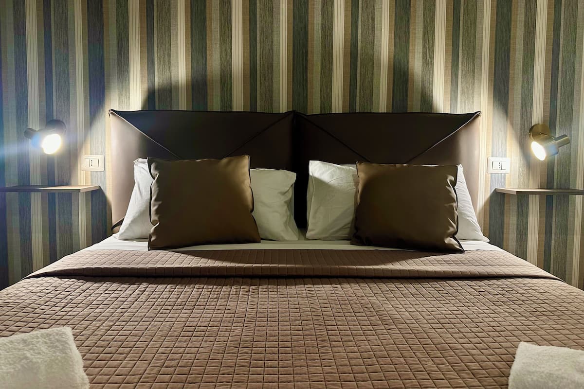 A neatly made bed with a brown quilted bedspread, two pillows, and a headboard with a unique design. There are two wall-mounted light fixtures on either side of the bed.