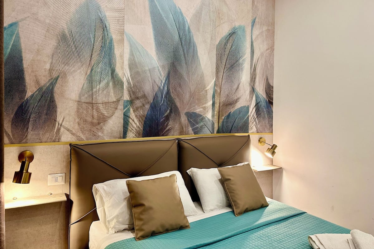 A bedroom with a bed, pillows, a headboard, a wall-mounted light, and a wall art piece.