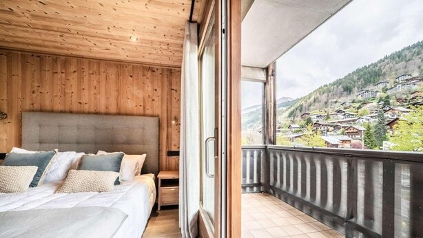 A bedroom with a wooden ceiling and a large bed, a small bedside table, and a view of a village through a balcony.