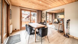 A modern dining area with a wooden table and chairs, a clock on the wall, and a view of a wooden building outside.