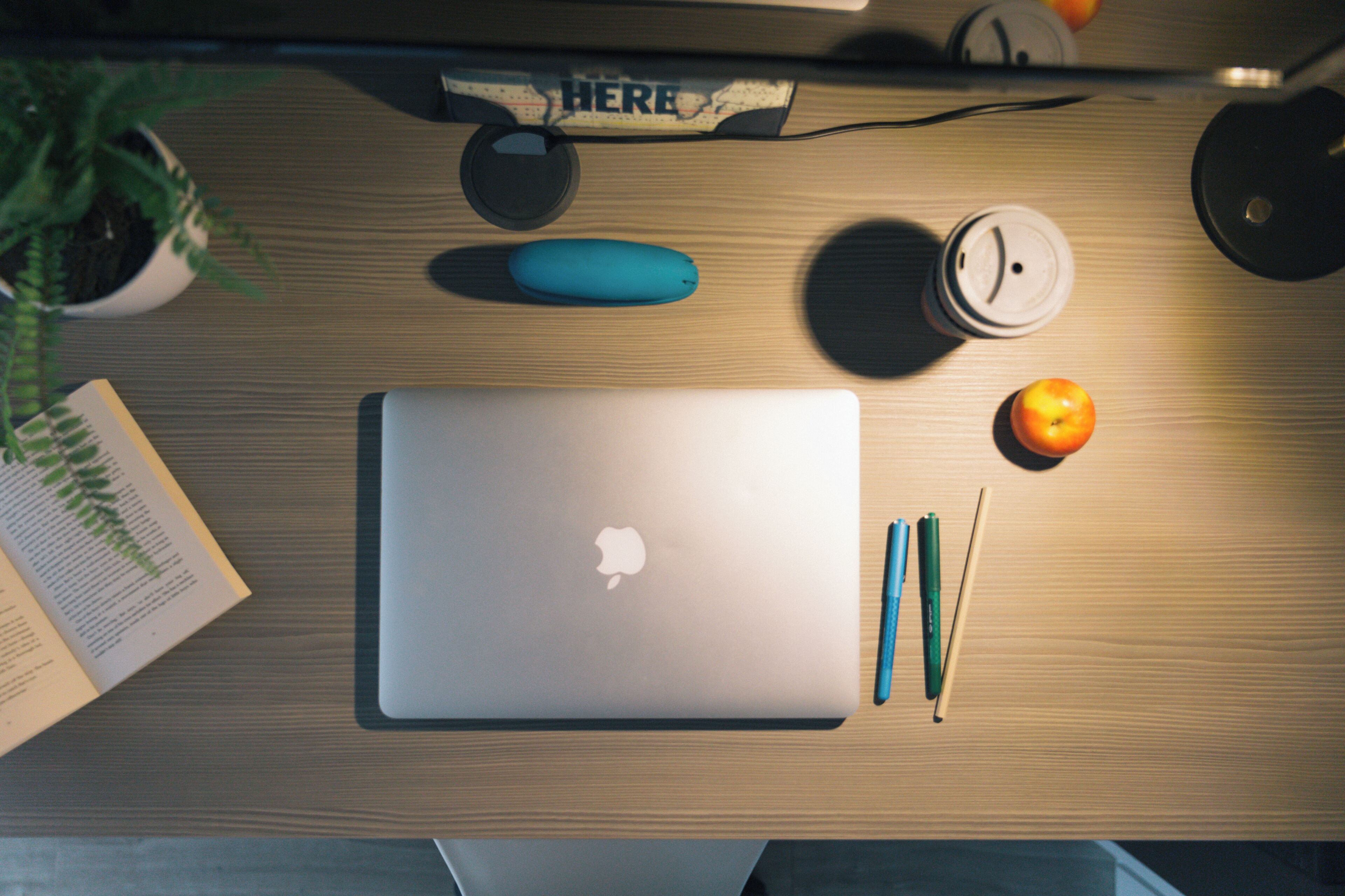 A laptop on a wooden desk with a book, a pen, a mouse, and a small orange.