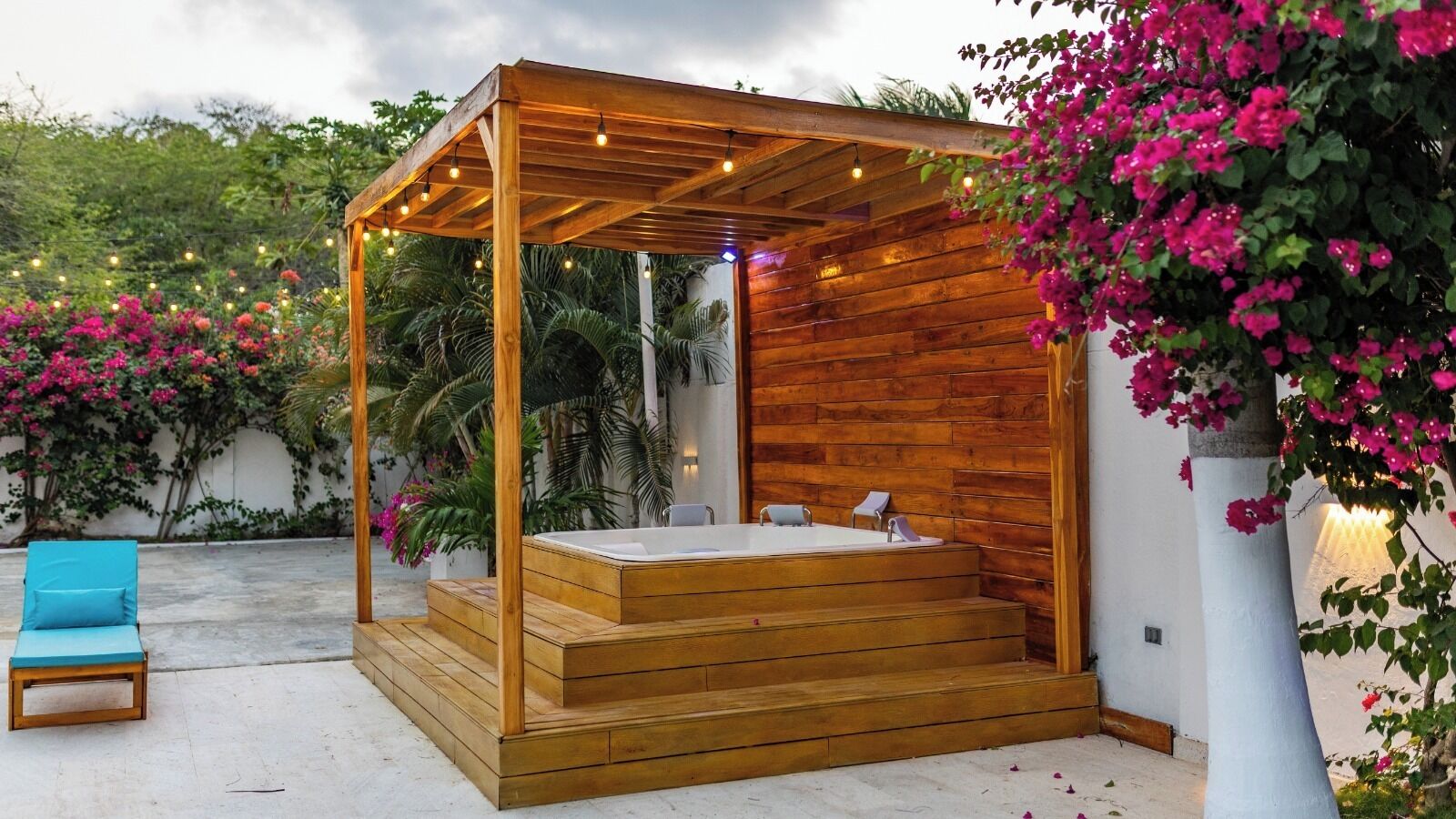A wooden outdoor hot tub with steps, surrounded by lush greenery and pink flowers.