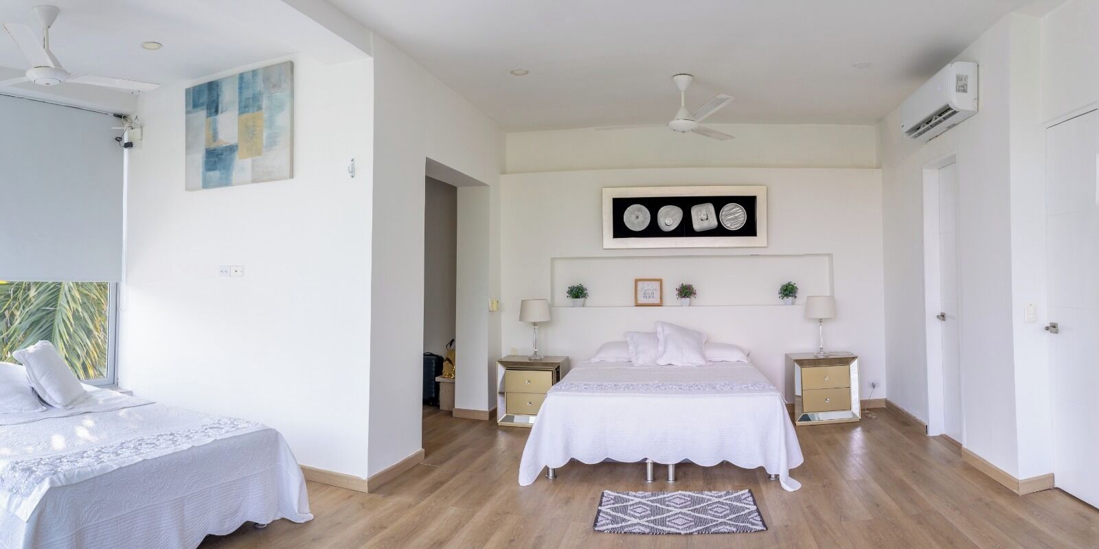 A bedroom with a bed, bedside tables, a ceiling fan, an air conditioning unit, and a window with a view of palm trees.