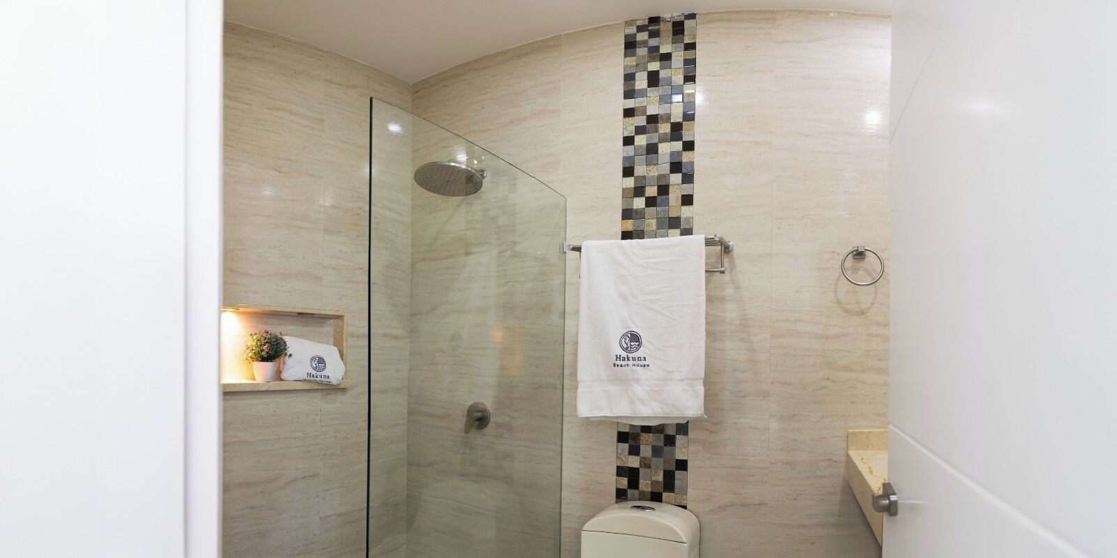 A modern bathroom with a glass shower enclosure, a toilet, and a sink. There is a towel rack with a towel hanging on it, and a small shelf with a plant.