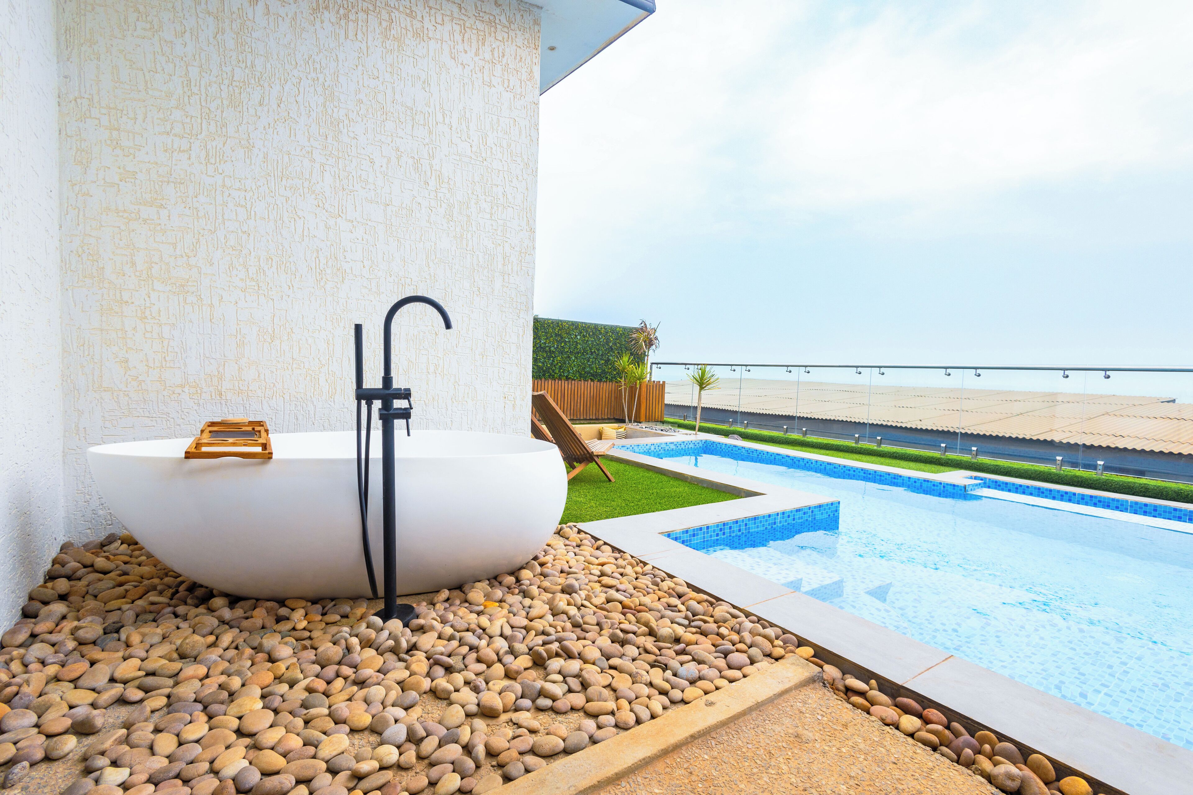 A modern outdoor bathroom with a freestanding bathtub, a shower, and a view of a pool and beach.