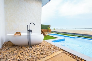 A modern outdoor bathroom with a freestanding bathtub, a shower, and a view of a pool and beach. - IAA Grande (Nyanyanu)