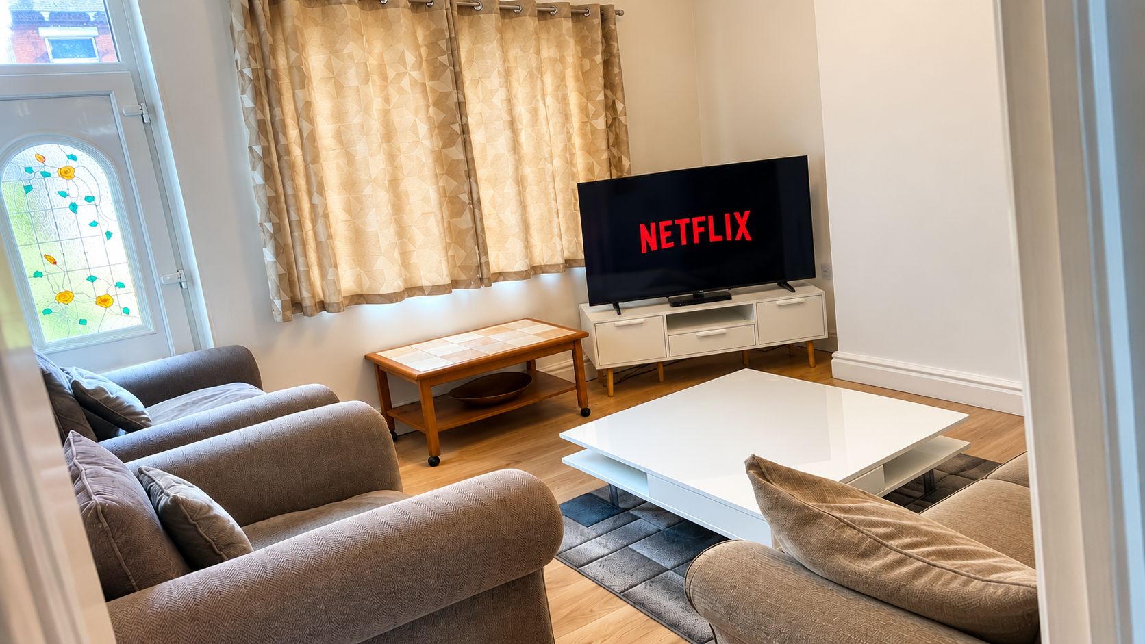 A living room with a flat-screen TV displaying 'NETFLIX,' a sofa set, a coffee table, and a window with curtains.