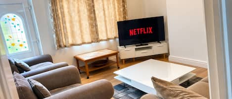 A living room with a flat-screen TV displaying 'NETFLIX,' a sofa set, a coffee table, and a window with curtains.