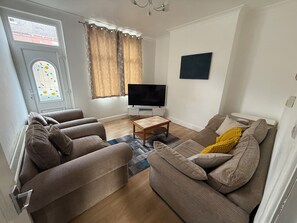 A living room with a sofa set, a TV, and a window with curtains. - 3BR-Sleep 10– Near Stadium,Centre & M-Links - Contractors / Corporate & Families (Leeds)