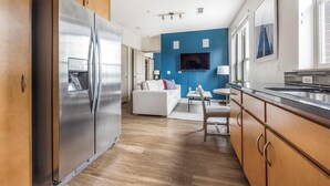 A modern kitchen with a stainless steel refrigerator, wooden cabinets, a dining area with a table and chairs, and a blue accent wall. - Landing | Amazing 1BD, Clubhouse, Gym (Denver)