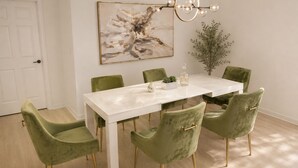 A dining area with a white table, green upholstered chairs, a pendant light, and a framed abstract painting.