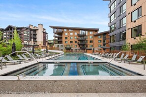 A swimming pool with lounge chairs and a modern building complex in the background. - Charlie's Slopeside: Ski-In/Out, Sleeps 7, Snowmass Base Village, Pool + Waterslide, Walk to Dining (Snowmass Village)