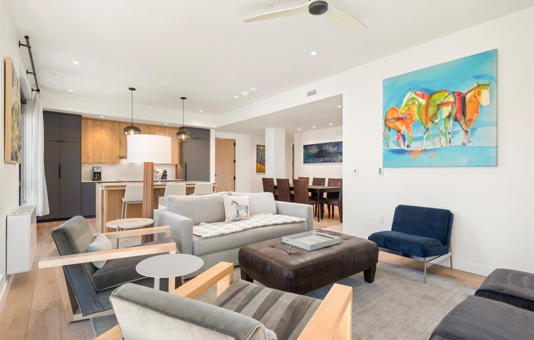 A modern living room with a sofa, ottoman, and chairs, a dining area with a table and chairs, and a kitchen with wooden cabinets and a dining table.