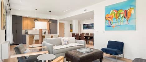 A modern living room with a sofa, ottoman, and chairs, a dining area with a table and chairs, and a kitchen with wooden cabinets and a dining table.