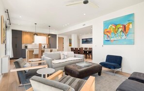 A modern living room with a sofa, ottoman, and chairs, a dining area with a table and chairs, and a kitchen with wooden cabinets and a dining table.