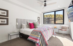 A bedroom with a bed, a nightstand, a chair, a window with a view of trees, and a ceiling fan.