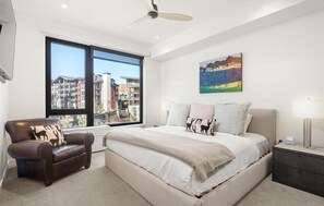 A modern bedroom with a large bed, a leather armchair, a nightstand, and a city view through a large window. - Charlie's Slopeside: Ski-In/Out, Sleeps 7, Snowmass Base Village, Pool + Waterslide, Walk to Dining (Snowmass Village)