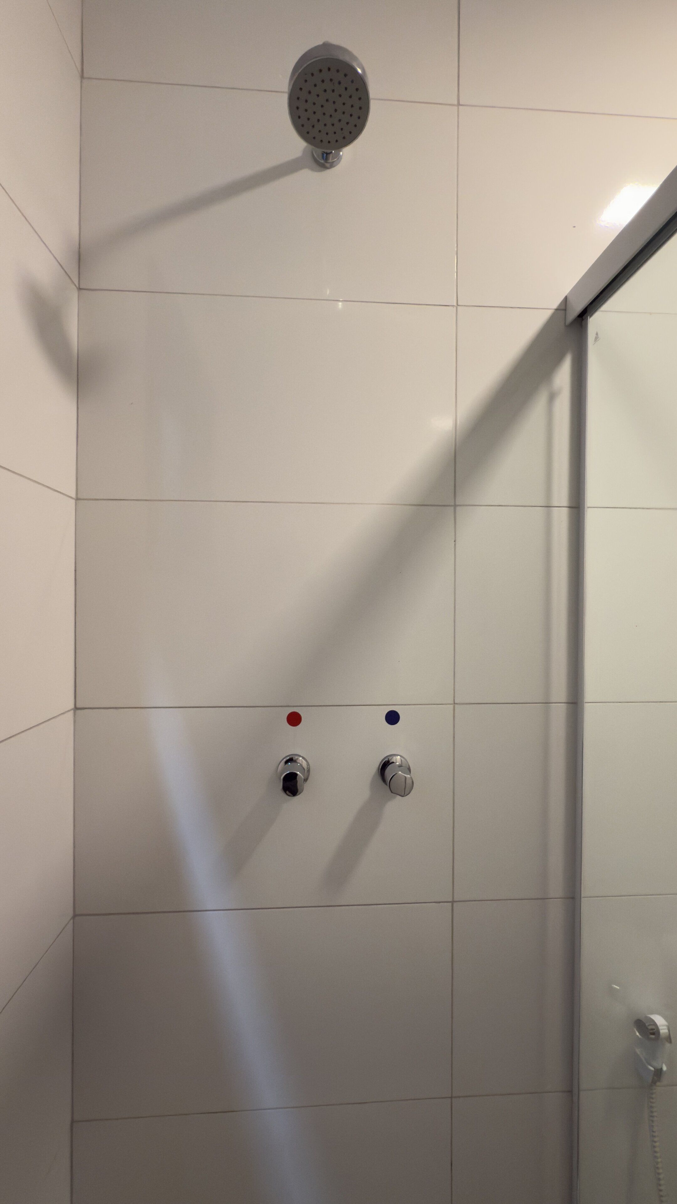 Chuveiro com chuveiro manual e duas manivelas, uma vermelha e outra azul, em parede revestida com azulejos.