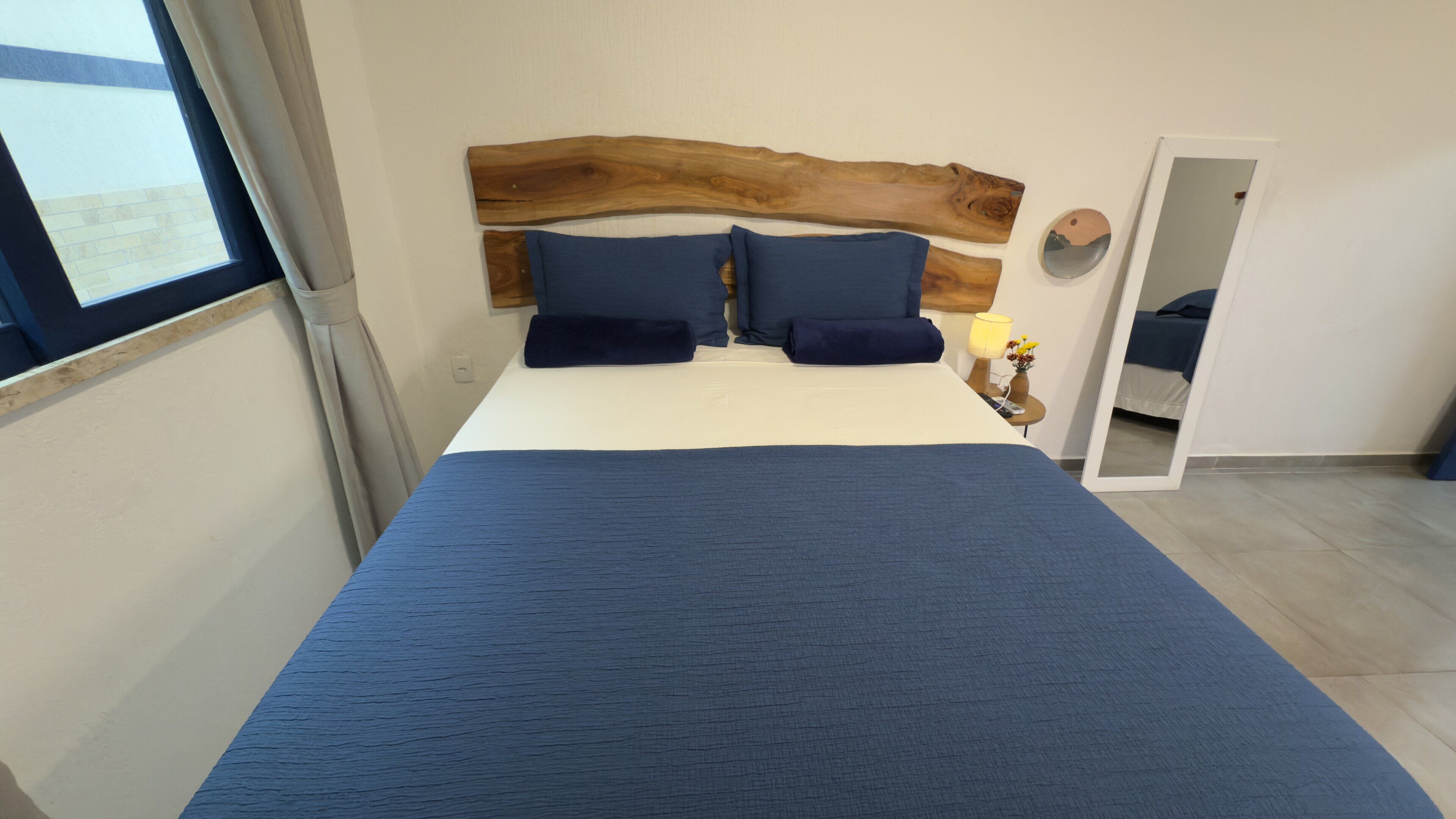 A bedroom with a large bed, a wooden headboard, two pillows, a bedside table with a lamp, and a mirror.