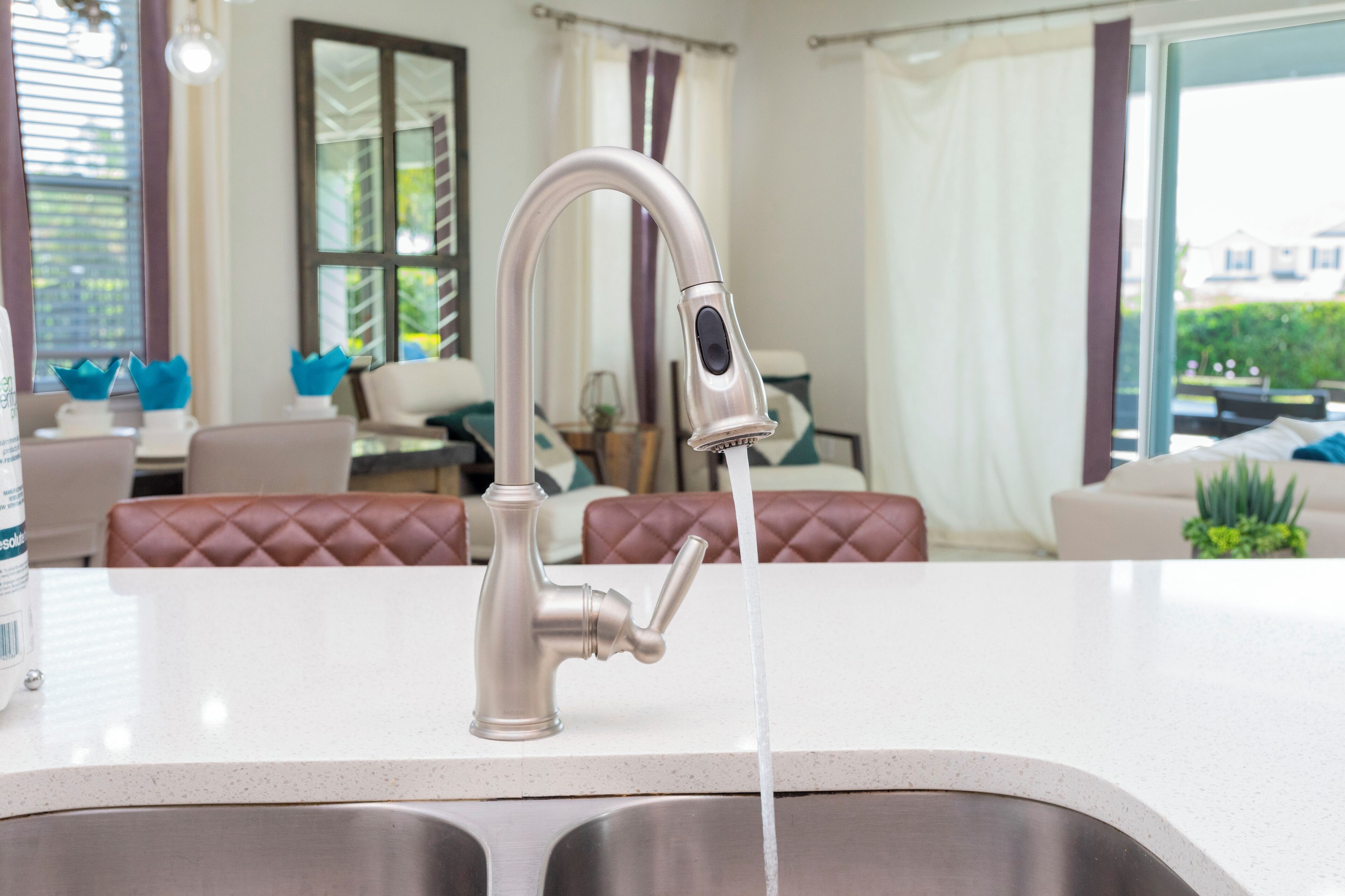 A modern kitchen with a stainless steel sink and faucet, a dining area with a table and chairs, and a view of a garden through a window.