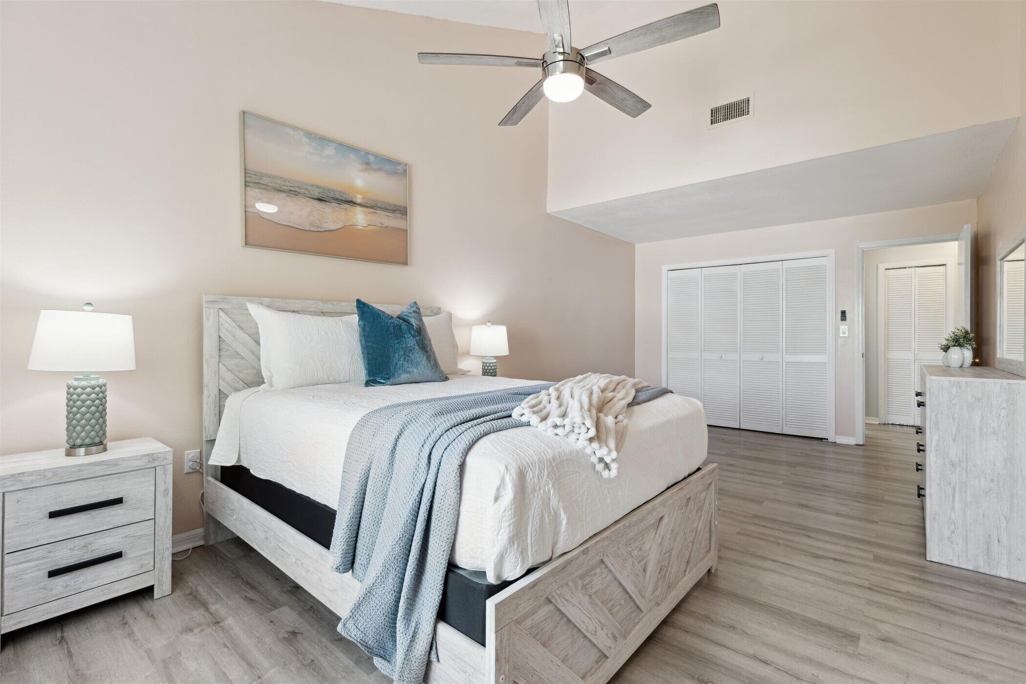 A bedroom with a bed, bedside tables, a ceiling fan, a painting, and a wardrobe.