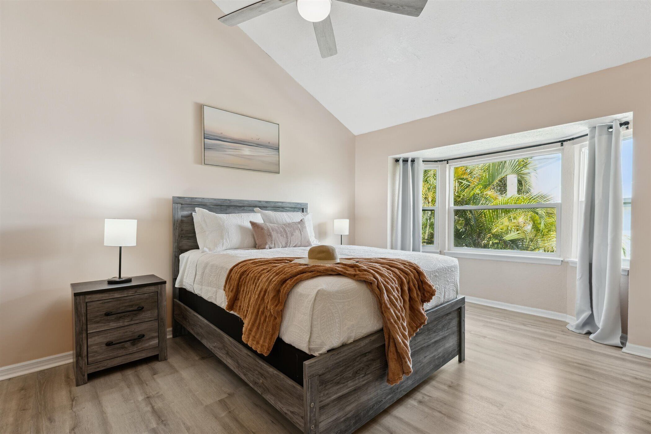 A bedroom with a bed, nightstand, ceiling fan, and large window with curtains.