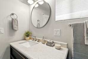 Baño con lavamanos doble, espejo redondo, toallero, planta en maceta y ventana con persianas.