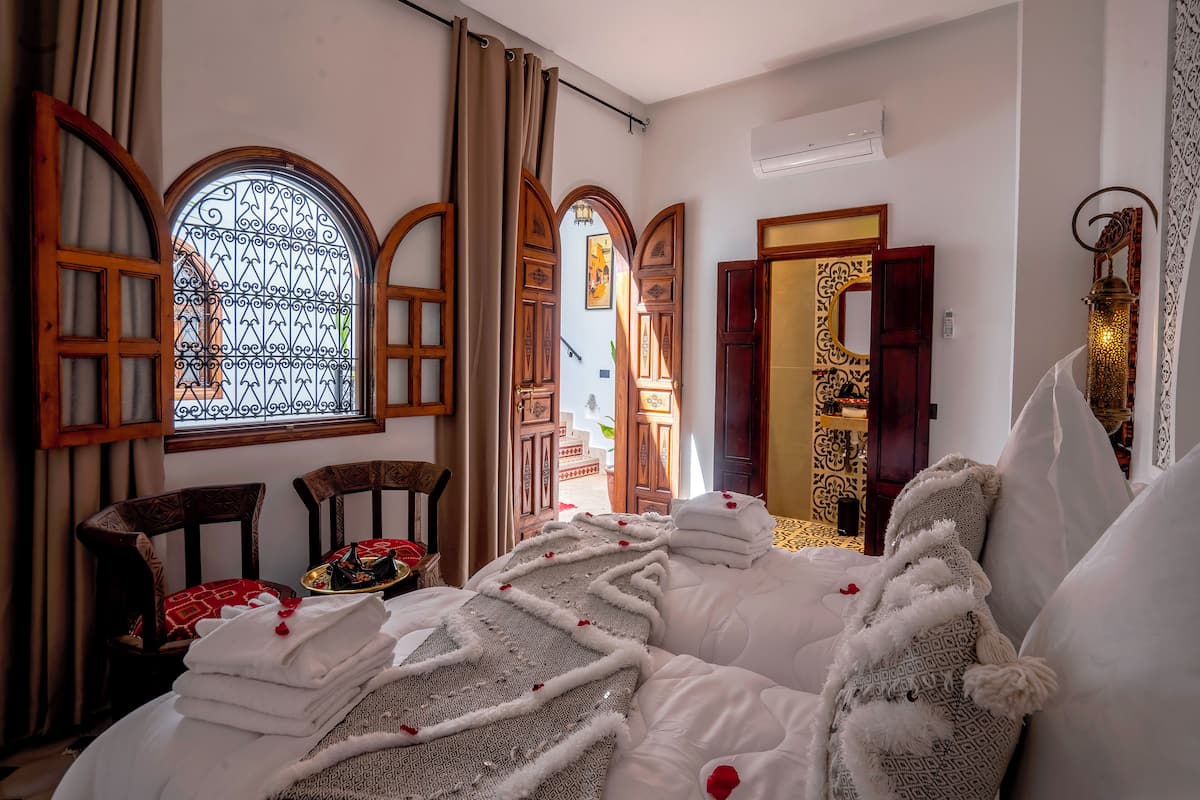 A room with a bed, two chairs, a window with curtains, and a door with a mirror.