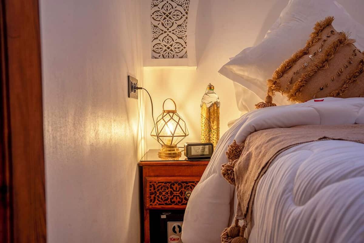 A neatly made bed with white bedding and a decorative pillow. A bedside table with a lamp and a decorative bottle is visible.