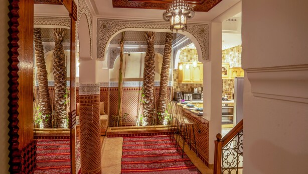 Un escalier orné de tapis rouges à motifs complexes et doté de rampes en bois, menant à une cuisine avec des meubles en bois et un lustre.