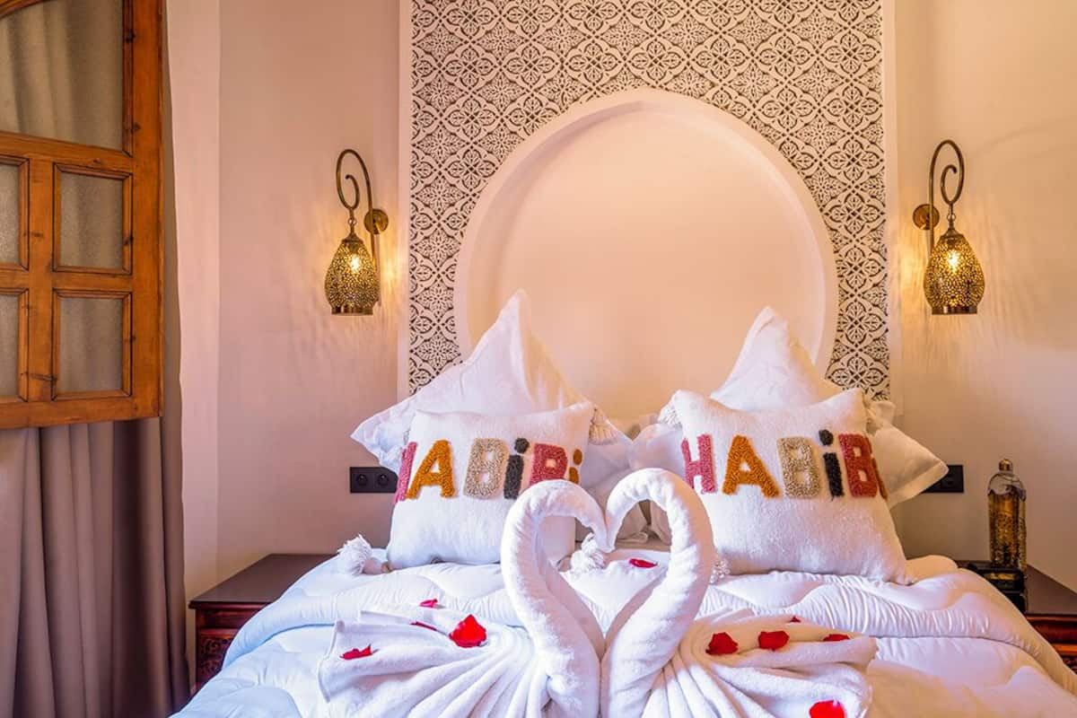 A hotel room with a bed adorned with white towels shaped like swans, pillows with the name 'JABIR' in colorful letters, and a patterned wall behind the bed.