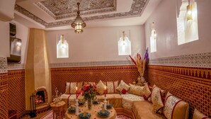 Un intérieur marocain traditionnel, avec un mur orné de motifs, un ensemble de canapés, un coin repas avec une table dressée pour quatre personnes et une lampe décorative suspendue.