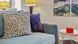 Un salon moderne avec un canapé bleu clair, un coussin à motifs, un objet rouge posé sur une table d’appoint et un tableau représentant un paysage accroché au mur.