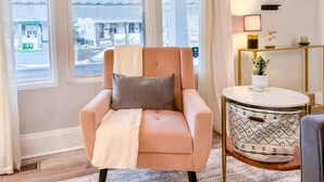 Une pièce avec un fauteuil de couleur pêche, une petite table ronde surmontée d’un coussin à motifs, et une vue sur la rue grâce à de grandes fenêtres.