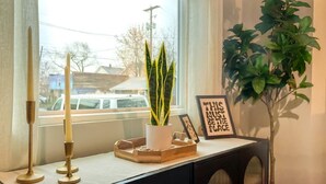Une pièce avec une fenêtre, une plante en pot, un meuble contenant des objets décoratifs et un panneau encadré.