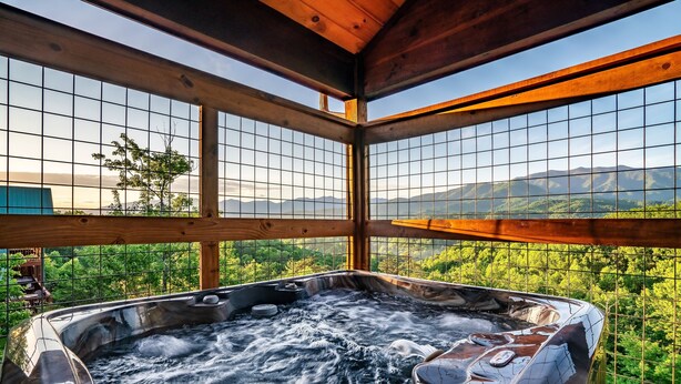 A hot tub with a view of mountains and greenery through a wooden structure with grid-patterned windows. - Rustic 3bed Cabin w/ Perfect Views of The Smokies! (Gatlinburg)