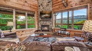 A cozy cabin interior with a stone fireplace, wooden walls, large windows, and comfortable seating. - Rustic 3bed Cabin w/ Perfect Views of The Smokies! (Gatlinburg)