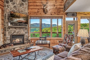 A cozy cabin interior with a stone fireplace, a mounted deer head, and a view of mountains through large windows. - Rustic 3bed Cabin w/ Perfect Views of The Smokies! (Gatlinburg)