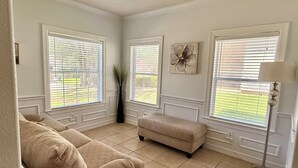 A living room with beige sofas, a coffee table, and a floor lamp. There are large windows with blinds, a vase with plants, and a wall art piece. - Charming 3-bedroom house in Lake Conroe, community pool and gym. (Montgomery)