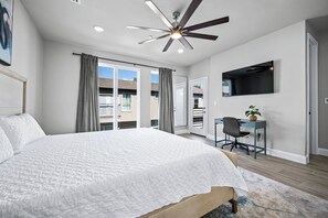 A bedroom with a large bed, a desk, a chair, a ceiling fan, and a TV mounted on the wall.