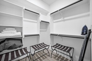 A compact room with a bed, two small tables, a bench, a shelf with folded towels, and a vacuum cleaner.