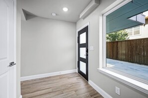 A hallway with a black door, a window with a view of a backyard, and a light switch on the wall.