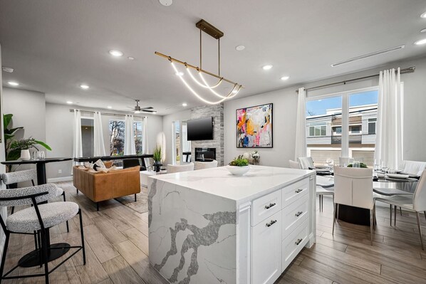 A modern kitchen with a marble island, a dining area with a white table and chairs, and a large window with curtains.